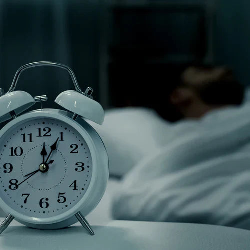 Alarm clock on a bedside table with a person sleeping in the background, representing the use of peptides for improving sleep and recovery